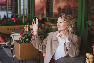 Beautiful business woman sitting in a street cafe and waving her hand, greeting someone