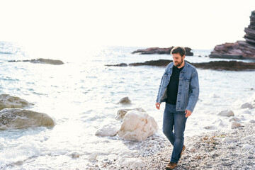 Pensive man in jeans and a denim jacket walks along a pebble beach near the water looking under his feet against a background of rocks - Powered by Adobe