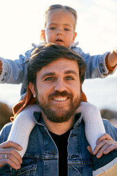 Portrait Of A Smiling Dad With A Little Serious Girl Spread Her Arms Out To The Sides On His Shoulders. Close-up