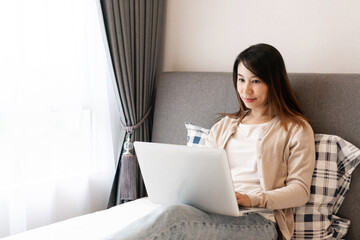 Happy casual beautiful Asian woman working on a computer laptop sitting on the bed in the house. Technology and lifestyle concept.