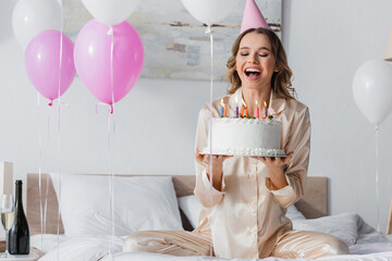 Happy woman in party cap holding birthday cake near balloons and champagne in bedroom