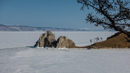 A two-headed rock, devoid of vegetation, rises above the frozen lake, and tiny silhouettes of people can be seen on the ice. Bare branches of a tree on a background of blue sky. Winter day. Baikal