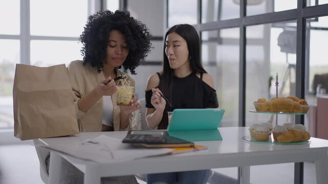 Two positive smiling young women eating noodles sitting at table in home office. Portrait of slim beautiful African American and Asian roommates having lunch break on remote working