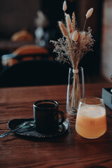 A cup of black coffee, orange juice and dried flowers in a glass vase standing on a wooden table in the interior of a cafe