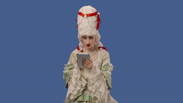 Portrait of courtier lady in white vintage lace dress and wig is viewing information on a digital tablet. Young woman posing in studio with blue screen background. Close up. Slow motion ready 59.94fps