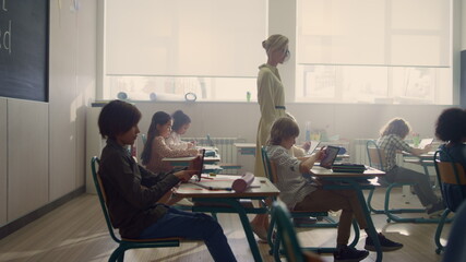 Schoolteacher walking in classroom with students. Pupils doing test on tablets