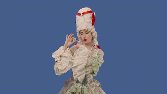 Portrait of courtier lady in vintage dress and wig showing a bitcoin coin and making thumbs up gesture. Young woman posing in studio with blue screen background. Close up. Slow motion ready 59.94fps.
