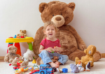 Sweet happy child boy having fun playing with his giant teddy bear and many colorful toys, indoor at home