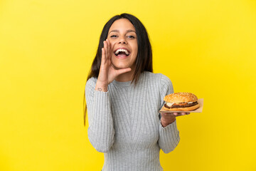 Young caucasian woman holding a burger isolated on yellow background shouting with mouth wide open