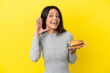 Young caucasian woman holding a burger isolated on yellow background listening to something by putting hand on the ear