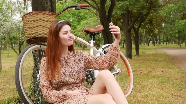 A young woman sits on the grass in a park in the city. bike for hire, rest outdoor. The girl uses her phone to video communicate with friends, photographed and takes a selfie