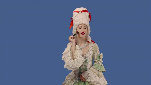 Portrait of courtier lady in vintage dress and wig is applying powder to her face using a makeup brush. Young woman posing in studio with blue screen background. Close up. Slow motion ready 59.94fps.