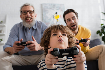 KYIV, UKRAINE - APRIL 12, 2021: Low angle view of excited child playing video game near father and granddad on blurred background