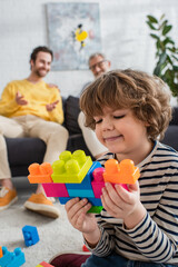 Obraz na płótnie Canvas Smiling boy playing building blocks near father and grandfather on blurred background