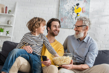 Smiling father with remote controller and grandfather with popcorn near child on couch