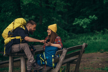 Plakat A couple of hikers sitting on a bench in the forest and making a break while using a smartphone