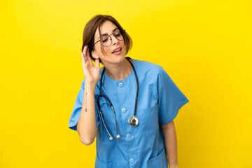 Surgeon doctor woman isolated on yellow background listening to something by putting hand on the ear