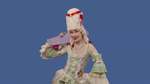 Portrait of courtier lady in vintage dress and wig, holding a skateboard and showing thumbs up gesture. Young woman posing in studio with blue screen background. Close up. Slow motion ready 59.94fps.
