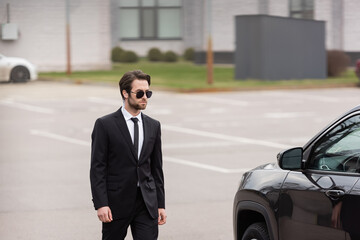 bearded bodyguard in suit and sunglasses with security earpiece walking near modern auto