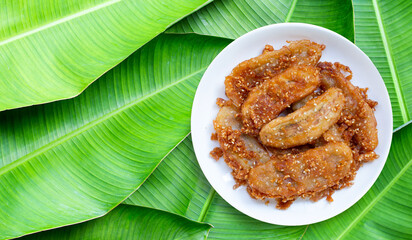 Fried bananas with sesame seeds on white ceramic round plate on banana leaves background. Top view