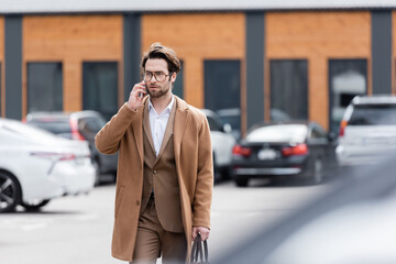 confident man in glasses and beige coat talking on mobile phone in outdoor parking