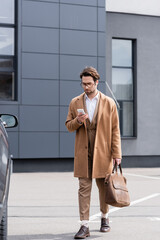 young man in glasses and coat standing with briefcase and texting on smartphone near car