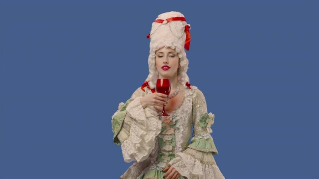 Portrait of courtier lady in vintage lace dress and wig, enjoying delicious wine or water in red glass. Young woman posing in studio with blue screen background. Close up. Slow motion ready 59.94fps.