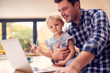 Working Father Using Laptop At Home On Kitchen Counter Whilst Looking After Young Daughter