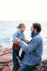 Laughing dad and little daughter sit on stones face to face near the sea, holding hands