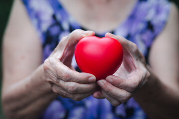 Hand of a senior woman holding a red heart shape while standing outdoors. Close-up photo. Aged people and healthcare concept