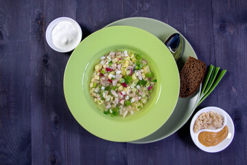 Okroshka,russian cold soup with meat, vegetables, kvass, sour cream, mustard, horseradish in green bowl with bread on black background
