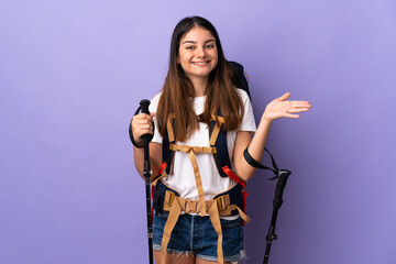 Young woman with backpack and trekking poles isolated on purple background extending hands to the side for inviting to come