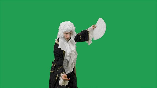 Portrait of courtier gentleman in black historical vintage suit and white wig, waving a fan and curtsy. Young man posing in studio with green screen background. Slow motion.