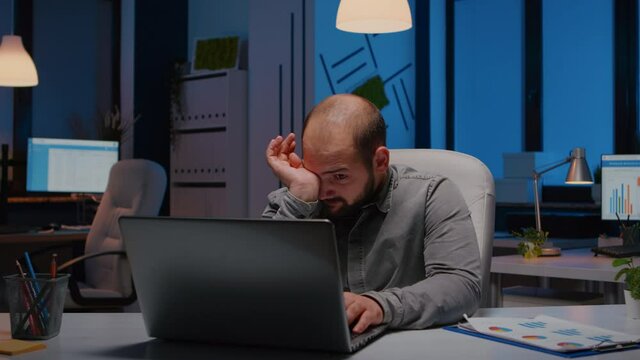 Exhausted sleppy businessman yawning while working at management project deadline late at night in start-up company office. Workaholic stressed entrepreneur typing financial strategy on laptop