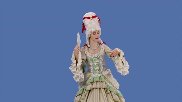 Portrait of courtier lady in white vintage lace dress and wig is waving a fan. Young woman posing in studio with blue screen background. Slow motion.