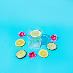 Lemons and cucumbers with a glass of water and flowers around. Blue background.