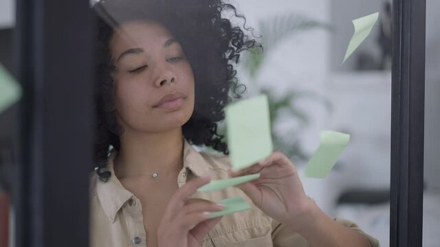 Portrait of serious satisfied young woman taking off sticky notes from glass door in home office. Confident charming African American businesswoman checking plan fulfilment indoors