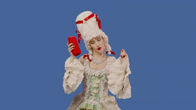 Portrait of courtier lady in white vintage lace dress and wig, listening to music with bluetooth portable speaker and dancing. Young woman posing in studio with blue screen background. Slow motion.