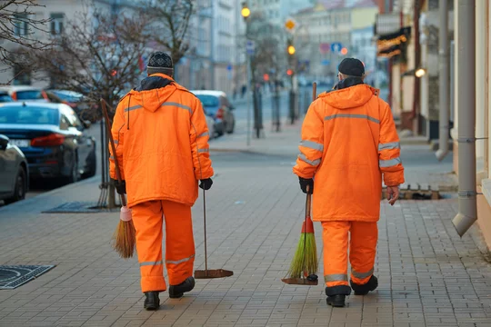 Fotka „Street sweaper clean road and pedestrian zone in the city. Municipal  workers sweep city street, janitors with broomstick and scoop for garbage.  Men in orange uniform collecting garbage from sidewalk“ ze
