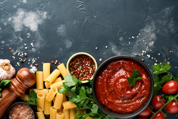 Pasta background. Pasta rigatoni, tomato ketchup sauce, olive oil, spices, parsley, and fresh tomatoes on a dark slate, stone or concrete background. Top view with copy space.