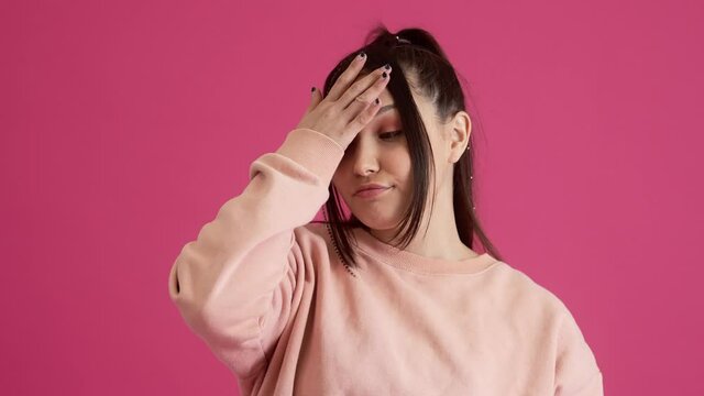 A confused young teenager girl is doing face palm standing isolated over pink wall in the studio