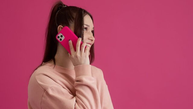 A pleased young teenager girl is talking on her smartphone standing isolated over pink wall in the studio