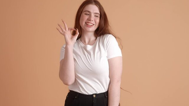 A confident plus size red-head woman is doing ok gesture while standing isolated over a beige wall in the studio
