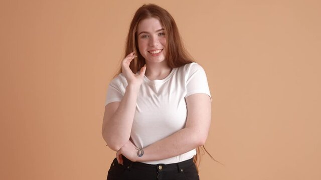 A happy plus size red-head woman is standing isolated over a beige wall in the studio