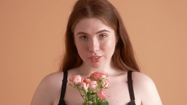 A close-up view of a beautiful plus size red-head woman is posing with flowers standing isolated over a beige wall in the studio