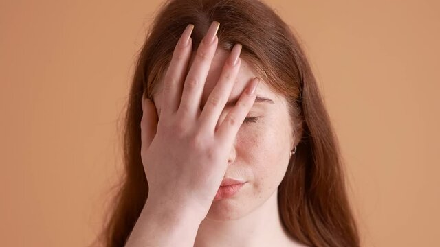 A close-up view of a displeased plus size red-head woman is doing face-palm gesture standing isolated over a beige wall in the studio
