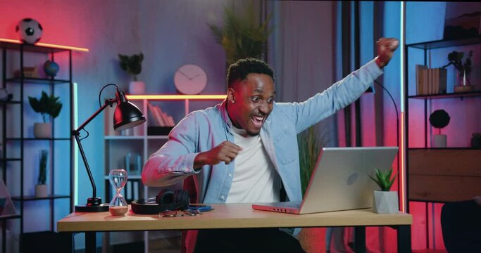Good-looking cheerful excited young african american sitting at the desk in front of laptop in night lighting room and rejoycing with funny reaction when get good news on screen