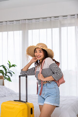 Obraz premium Happy young Asian woman tourist standing with luggage and backpack in bedroom, ready for vacation travel trip, looking at camera.