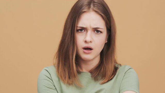 A displeased young woman is looking to the camera standing isolated over a beige wall in the studio