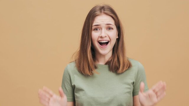 A happy young woman is looking to the camera while clapping her hands standing isolated over a beige wall in the studio